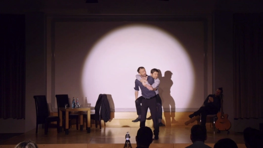 Zwei Schauspielende für Improvisationstheater im Scheinwerferlicht auf einer Bühne. Eine Person sitzt im Huckepack auf dem Rücken der anderen Person.