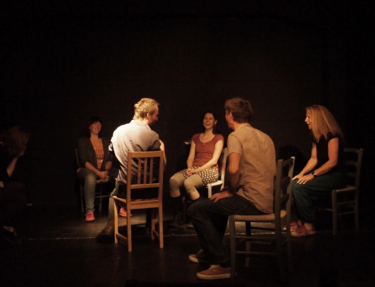 Die Teilnehmenden eines Improtheater-Workshops sitzen auf Stühlen und schauen sich an.